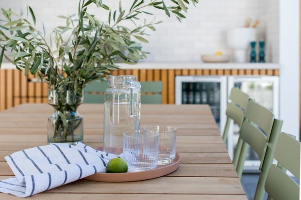 A bright, contemporary dining area centered on a long light timber dining table. The table is styled with a large leafy branch arrangement, a ceramic tray, glass water jugs and drinking glasses, a lime, and a striped linen napkin. Pale green dining chairs line the right side. In the background, a white tiled or brick-look wall, timber slat detailing, and a compact drinks cabinet or refrigerator are visible.