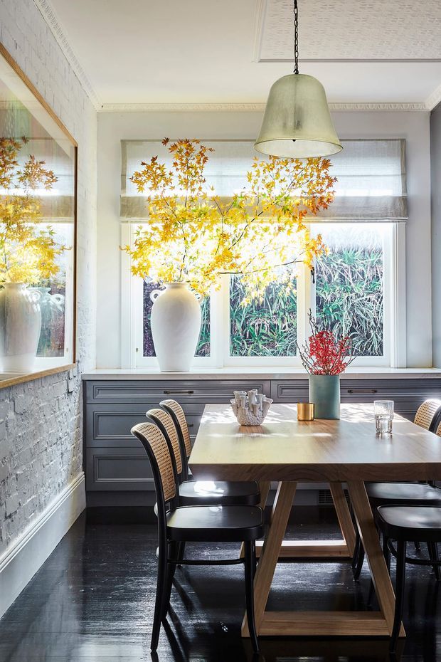 A bright, refined dining room with a dark timber dining table and woven-back black chairs positioned beside large windows. Grey-blue built-in cabinetry runs beneath the windows, while a large ceramic vase with autumnal branches forms a focal point. The room features white-painted textured walls, ornate ceiling cornices, a decorative ceiling panel, dark timber flooring, a large framed mirror, and a suspended aged-metal pendant light.