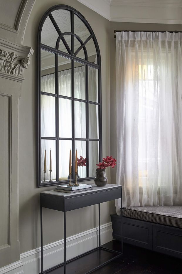 A refined traditional interior corner featuring a tall arched black-framed mirror, ornate classical wall mouldings and corbels, sheer white curtains, and a slim black console table styled with books, candlesticks, and a vase of flowers. A built-in dark bench seat sits beneath the window, creating an elegant, softly lit transitional space.