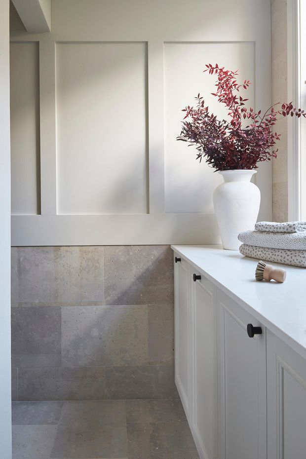 Bright, refined laundry room with white raised-panel cabinetry, a pale white benchtop, and large grey-beige stone tiles wrapping the lower wall and floor. A decorative white vase with burgundy foliage, folded towels, and a wooden brush add understated styling. The room has a calm, traditional-inspired fit-out with natural light from the right.
