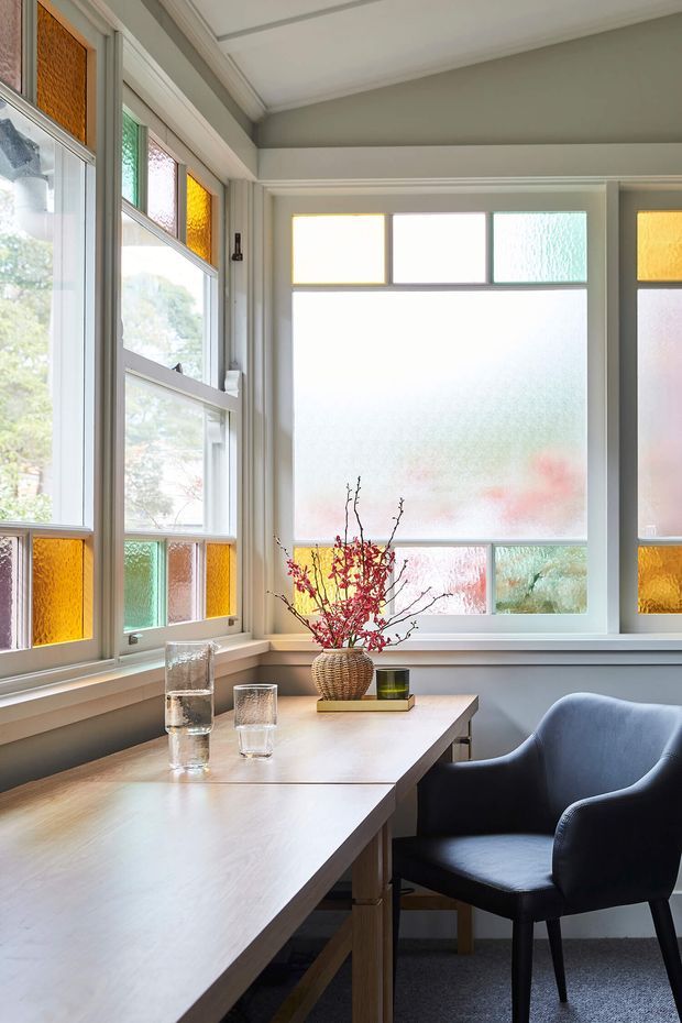 A bright, compact dining nook with a pale timber dining table, upholstered dark armchairs, and a woven vase with branches. The room is defined by extensive leadlight-style stained and textured glass window panels in amber, green, pink, and blue, set within white-painted frames. Soft grey walls, a simple coffered ceiling, and carpet create a calm, refined atmosphere.