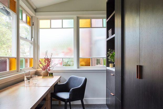 A compact, bright home office or study nook with a long light-wood desk running along the windows, a single dark upholstered armchair, and extensive frosted and coloured stained-glass-style window panels. Dark timber built-in shelving and storage occupy the right side, with a small plant and simple decorative objects. The room has a calm, refined contemporary character with strong natural light and contrasting dark joinery.