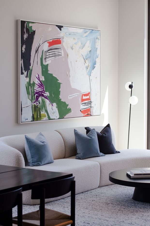 A bright, refined living room featuring a low-profile cream bouclé sectional sofa with blue and charcoal cushions, a large expressive abstract artwork in a pale frame, sculptural black-and-white floor lighting, a dark wood or black coffee table, and a textured neutral rug. Strong natural light enters from the right, creating a calm contemporary atmosphere.