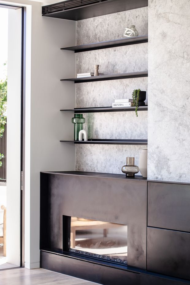 Close-up interior view of a contemporary built-in fireplace and dark custom cabinetry, with open floating display shelves against a textured grey feature wall. Neutral plaster walls, pale timber flooring, and restrained decorative objects create a refined, minimalist living-room feature.