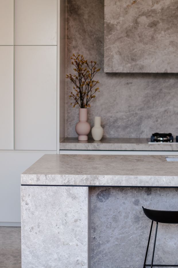 Close-up view of a refined contemporary kitchen featuring a large stone-clad island with a slim waterfall edge and seating. The island and rear counter use a mottled grey stone finish, complemented by pale flat-panel cabinetry and a textured grey feature wall with a floating artwork panel. Minimal decorative vases and a black bar stool add restrained contrast.