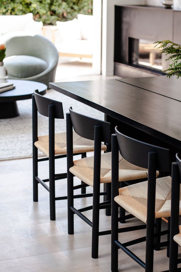Bright contemporary dining and living area featuring a long dark dining table with black timber-framed chairs and woven natural-fibre seats. A round coffee table, pale upholstered lounge chair, light tiled floor, large glazing, and a dark built-in fireplace or media surround are visible. The space has a clean, refined black-and-neutral palette with indoor greenery.