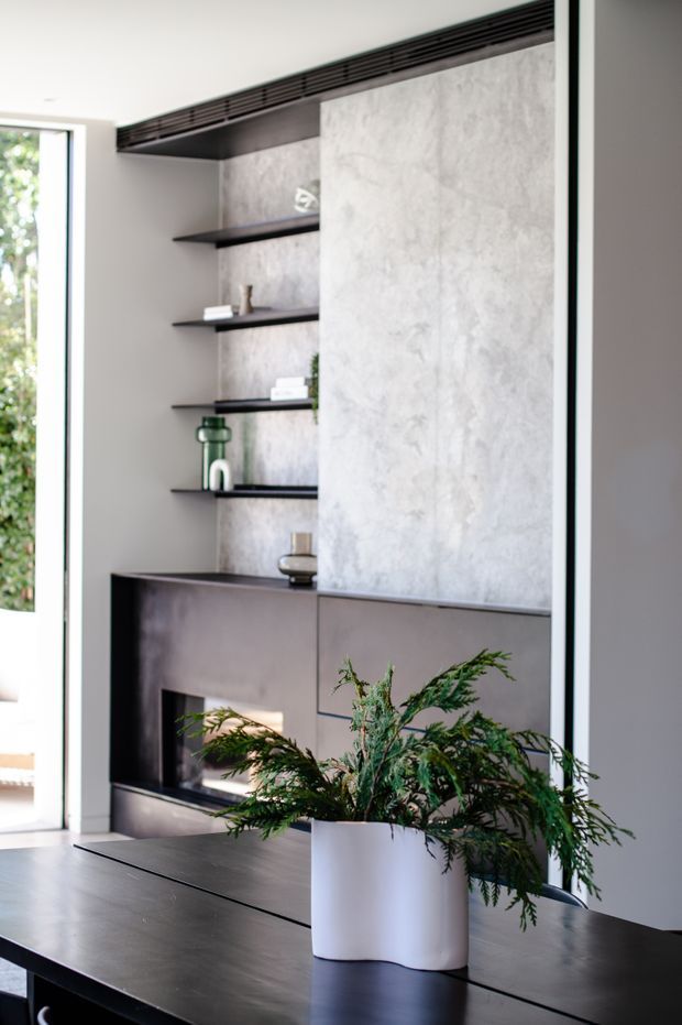 A refined contemporary living area featuring a dark built-in fireplace wall with integrated cabinetry and open display shelving. A large textured light-grey feature panel contrasts with dark timber or painted joinery, while a black stone-like console or table occupies the foreground with a potted fern. Full-height glazing at left brings in natural light.