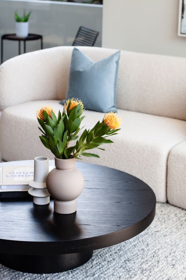 A softly styled living room vignette featuring a large curved cream boucle sofa, a pale blue cushion, and a low round black timber coffee table. The table holds a sculptural ceramic vase with orange flowers, a white candleholder, and a book. A textured light rug, minimalist side table, potted plant, and outdoor chair visible through the glazing complete the scene.