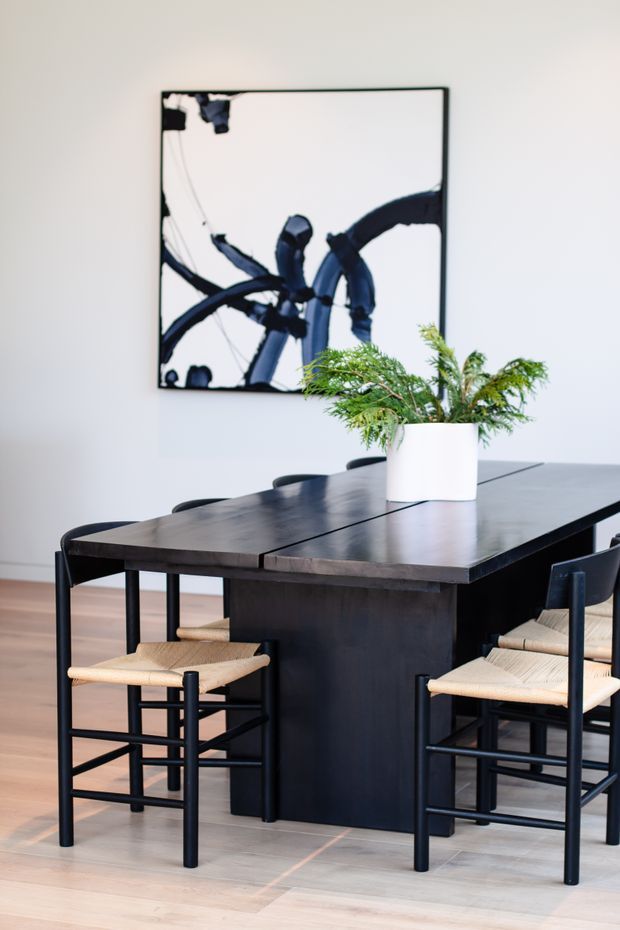 A refined dining area featuring a large dark timber dining table with a substantial central pedestal base, surrounded by black-framed woven-seat chairs. A white cylindrical planter with greenery sits on the table, while a large abstract black-and-white artwork hangs on the pale wall behind. Light timber flooring and a restrained monochromatic palette create a contemporary, calm interior.