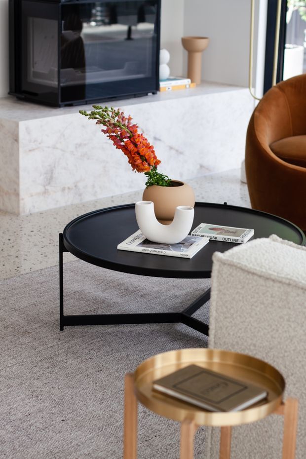 A close-up of a refined contemporary living room featuring a low black round coffee table with a thin metal frame, styled with books and a white sculptural vase containing orange-red foliage. A pale stone or marble fireplace surround forms the backdrop, with a dark television or display unit above it. The room includes a tan leather lounge chair, a light textured sofa, grey carpeting, and a partially visible brass-toned side table.