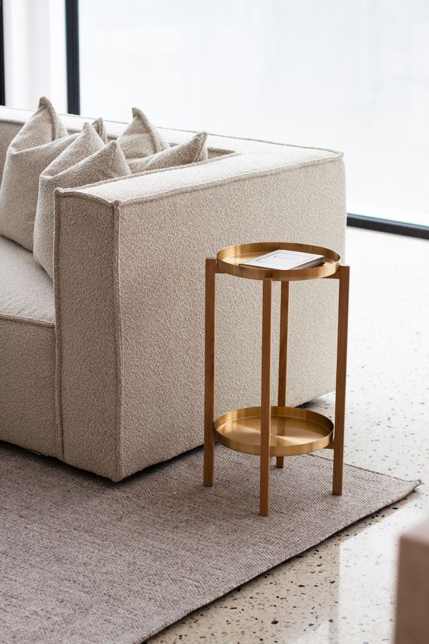 Bright contemporary interior detail featuring a plush cream boucle sofa with tailored square arms and cushions, a slim round two-tier brass-gold side table, a neutral textured area rug, and a polished terrazzo floor beside large dark-framed glazing.