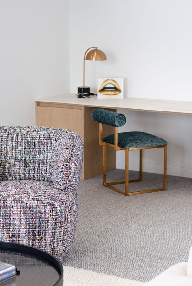 A softly styled contemporary interior corner featuring a light timber built-in desk or console with pale stone-like top, a brass dome desk lamp, teal upholstered cantilever chair with brass-toned frame, and a multicoloured textured armchair. Neutral painted walls and grey carpet create a calm, refined workspace or living area.