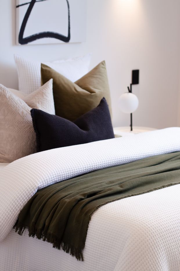 Close-up of a neatly styled bedroom featuring a white textured bedspread, layered cream, olive and deep navy cushions, and an olive throw. A minimalist black-and-white bedside lamp and abstract black line artwork sit against light neutral walls, creating a calm, refined atmosphere.