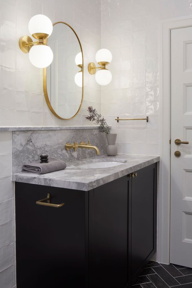 A refined bathroom vanity vignette featuring a dark built-in cabinet with brass hardware, a grey-veined stone benchtop and integrated basin, glossy white tiled walls, and a large oval brass-framed mirror. Globe wall sconces provide warm decorative lighting, while a white panelled door and dark geometric floor tiles complete the space.