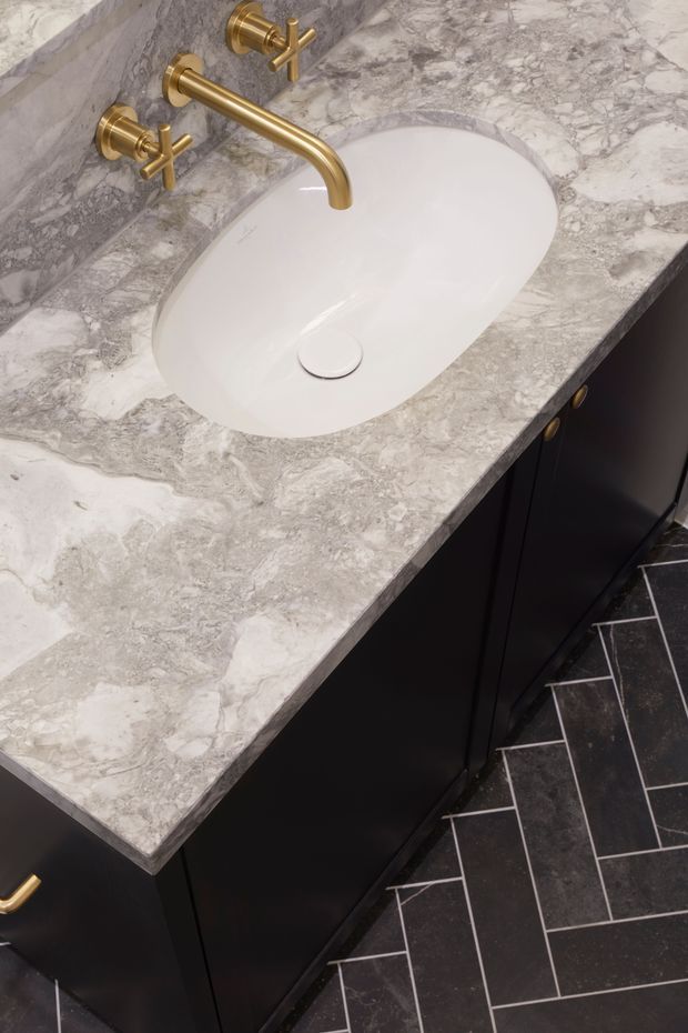 Close-up view of a refined bathroom vanity featuring a white oval undermount basin, grey-and-white natural stone benchtop and matching wall upstand, matte black cabinetry with brass handles, and a brushed brass wall-mounted tap. Dark rectangular floor tiles are laid in a herringbone pattern.