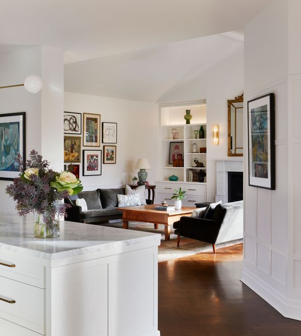 A bright, refined open-plan interior viewed from a white kitchen island, looking into a living room. The space features white panelled cabinetry, a pale stone benchtop, dark timber flooring, a fireplace with a marble surround, built-in shelving, framed artwork, a dark sofa and armchairs, and a timber coffee table. Abundant natural light and layered decorative styling create a warm, classic atmosphere.