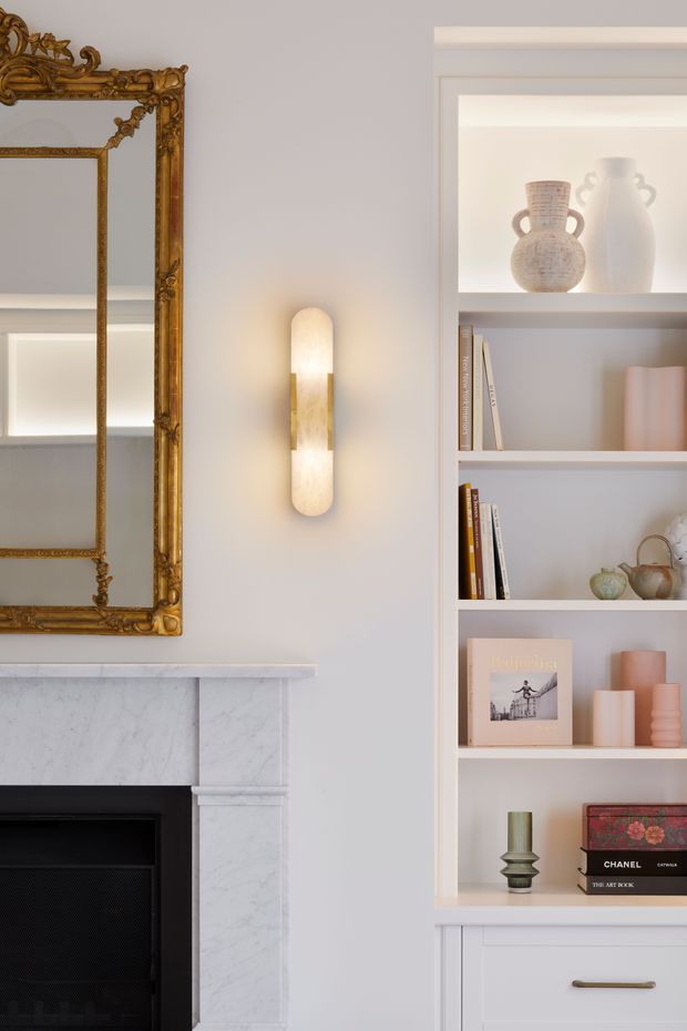 A refined living room vignette featuring a white marble fireplace with a black firebox, an ornate antique gold-framed mirror, a sculptural wall sconce, and custom white built-in shelving filled with books and decorative ceramics. The styling combines classic architectural details with soft contemporary furnishings and warm ambient lighting.