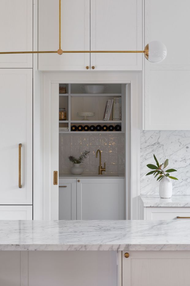 Detailed view of a refined white kitchen with raised-panel cabinetry, brass hardware and a linear brass-and-glass pendant light. A compact butler's pantry is visible through a central doorway, featuring open shelving, white cabinetry, a small sink with brass tapware, and glossy textured tile backsplash. White marble with grey veining is used for the main island and surrounding benchtops, creating a bright, classic interior.