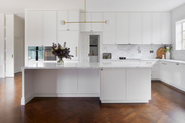 Bright, refined white kitchen with extensive full-height cabinetry, a marble-look backsplash and benchtops, a central island, brass pendant lighting and fittings, and warm dark timber herringbone flooring. The space includes a concealed pantry or appliance cupboard and opens toward an adjacent hallway or room.