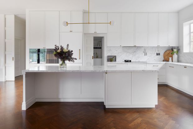 Bright, refined white kitchen with extensive built-in cabinetry, a large marble-look island, veined stone splashback, integrated appliances, and dark herringbone timber flooring. A brass linear pendant light and floral arrangement add understated warmth, while a window and mirrored surfaces bring in natural light.