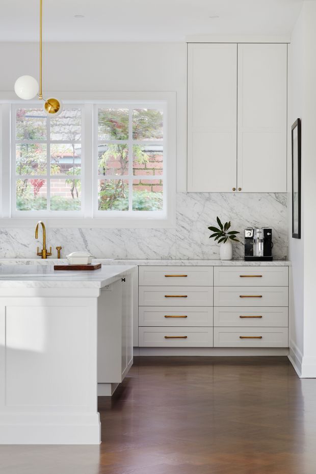 Bright, refined white kitchen with a central island, marble backsplash and benchtops, shaker-style cabinetry, brass hardware and tapware, timber flooring, and large divided-pane windows overlooking greenery. A small coffee station and potted plant sit on the rear counter, while a sculptural brass-and-white pendant adds a decorative focal point.
