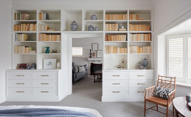 Bright, traditionally styled bedroom with extensive custom white built-in bookcases and drawer cabinetry framing an open doorway to a sitting room. The room includes neutral carpeting, a partially visible upholstered bed, a woven wood armchair with patterned cushions, and plantation shutters across the large window. The built-in shelving is filled with books and decorative objects.