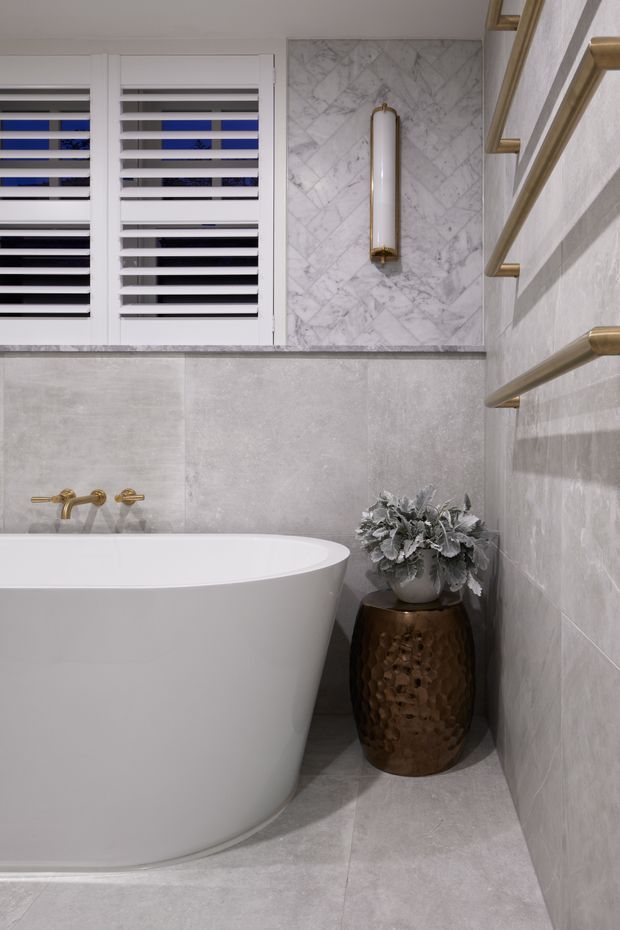 A refined contemporary bathroom featuring a freestanding white bathtub, large-format pale grey stone-look wall and floor tiles, a marble-look feature panel with a vertical brass wall light, white plantation shutters, brushed brass tapware and towel rails, and a hammered bronze planter stool with foliage.