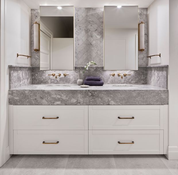 A refined double-vanity bathroom with white shaker-style cabinetry, a dramatic grey-veined stone benchtop and matching backsplash, paired rectangular mirrors, brass wall-mounted tapware and sconces, and a pale grey tiled floor.