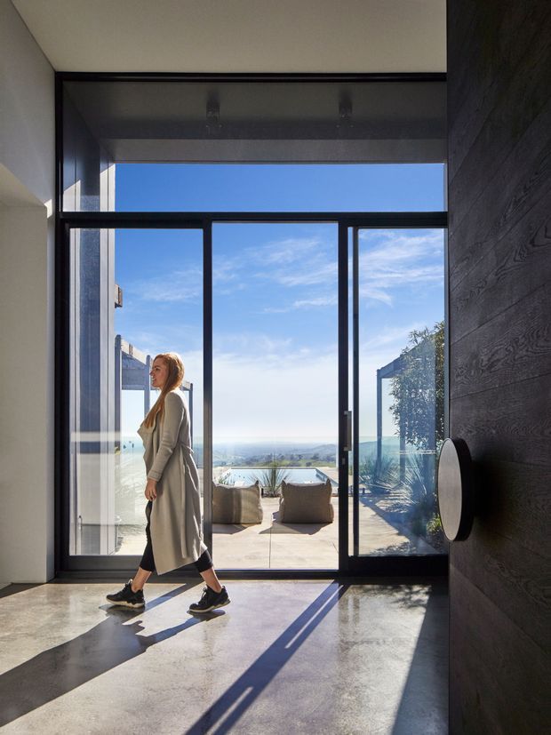 A bright, minimalist interior entryway with a polished concrete floor, dark timber wall panelling, and expansive black-framed floor-to-ceiling glazing opening onto a sunlit terrace. A person is walking through the space, with distant landscape and water visible beyond the outdoor area.