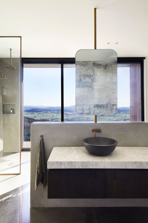 A refined contemporary bathroom vanity overlooking a rolling rural landscape. The room features a dark timber floating vanity, pale stone benchtop, sculptural dark vessel basin, aged brass wall-mounted tapware, a large mottled rectangular mirror, black-framed floor-to-ceiling glazing, and a textured pale plaster or concrete partition. A frameless glass shower is visible at left.