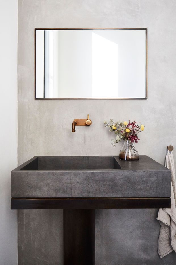 A minimalist bathroom vanity with a large rectangular mirror, a monolithic dark grey concrete or stone basin, exposed wall-mounted copper tapware, and a dark timber support cabinet. The textured light grey wall and restrained styling create a refined, earthy atmosphere.