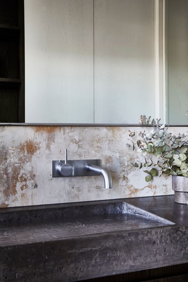 Close-up of a refined bathroom vanity featuring a dark integrated basin and countertop, a wall-mounted brushed metal tap, weathered textured stone or plaster splashback, pale wood-grain cabinetry, and a large mirror. A small potted leafy arrangement adds an organic accent.
