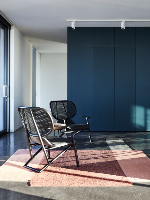 A minimalist contemporary interior, likely a living or lounge area, with two black mesh lounge chairs on a muted pink and burgundy geometric rug. A full-height dark navy built-in wall provides a strong backdrop, while expansive glazing on the left brings in bright natural light and casts long shadows across the polished grey floor. White track lighting and pale walls complete the restrained, modern space.