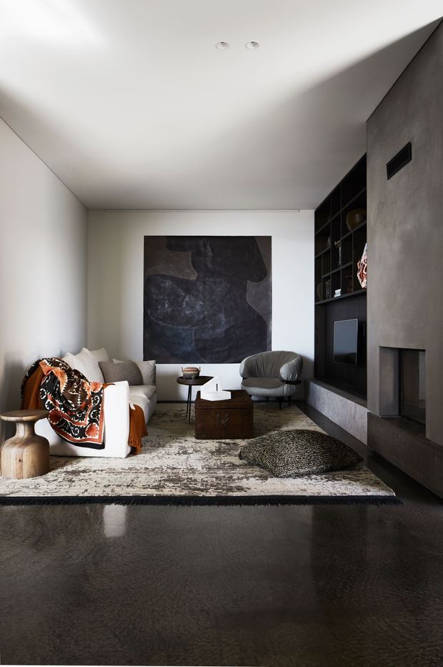 A refined contemporary living room with a minimalist built-in entertainment wall and fireplace. The space features white walls and ceiling, a dark textured feature wall, black open shelving, polished dark flooring, a large neutral patterned rug, sculptural lounge chairs, a white sofa with a bold patterned throw, and understated decorative objects.
