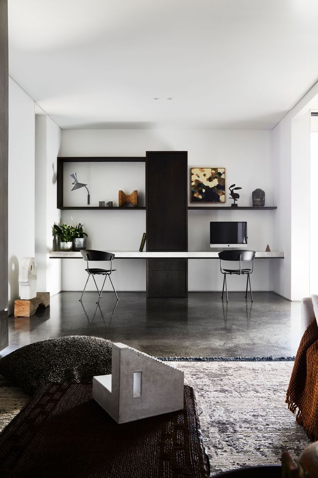 A bright, minimalist open-plan living and work area with a built-in wall-mounted desk, two black chairs, open shelving, dark timber cabinetry, a computer, artwork, and plants. The room has white walls and ceiling, polished dark concrete flooring, and layered textured rugs. A low concrete armchair or sculptural seat occupies the foreground, with a glimpse of upholstered seating at right.