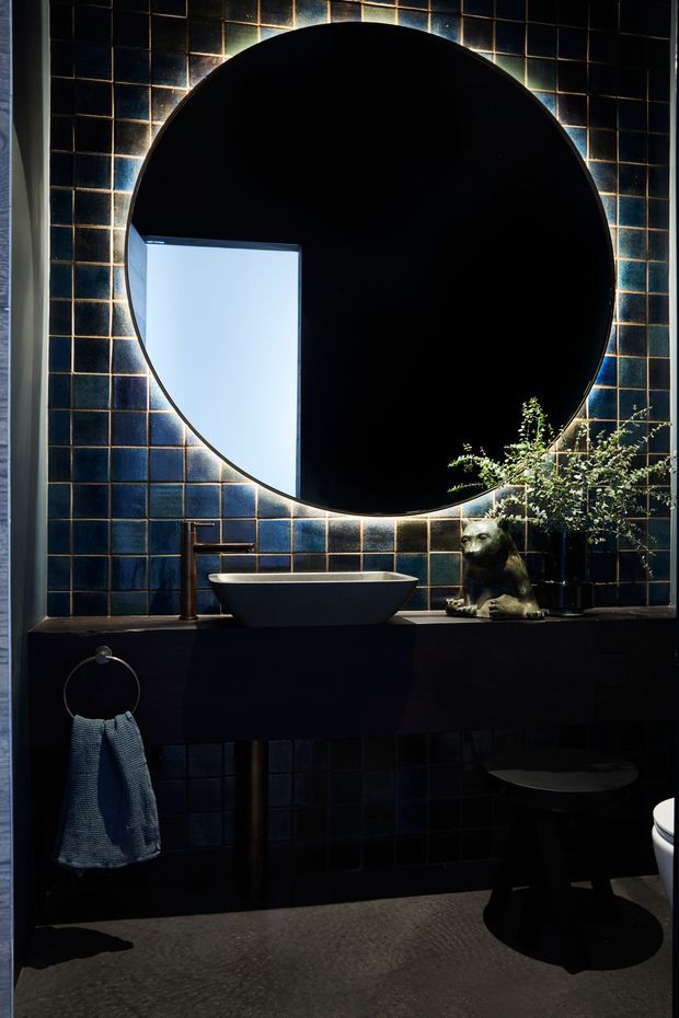 A dark, atmospheric bathroom featuring a large round black-framed mirror, deep blue-black square wall tiles with contrasting grout, a matte black floating vanity, vessel basin, dark tapware, and a small sculptural animal ornament with greenery. A towel ring and stool are visible, with dramatic daylight reflected in the mirror.