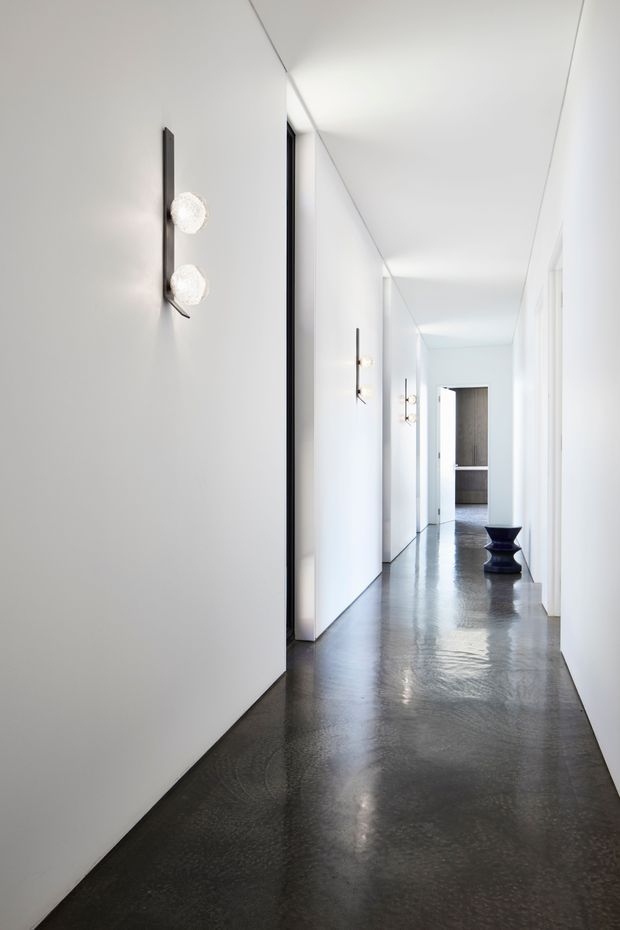 A long, minimalist interior hallway with crisp white walls and flush doors, a highly polished dark concrete floor, and sculptural wall sconces. The corridor leads toward an open doorway, with a small dark sculptural stool or pedestal positioned along the passage.