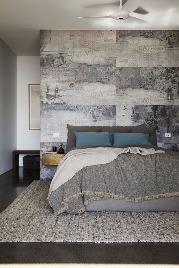 A contemporary bedroom with a low upholstered bed dressed in layered grey bedding and blue-grey cushions. The focal wall is covered in a large-scale monochromatic patterned mural or textured artwork, flanked by small wall lights. Dark flooring, a textured neutral rug, a wood-framed mirror, and a white ceiling fan complete the restrained, modern interior.