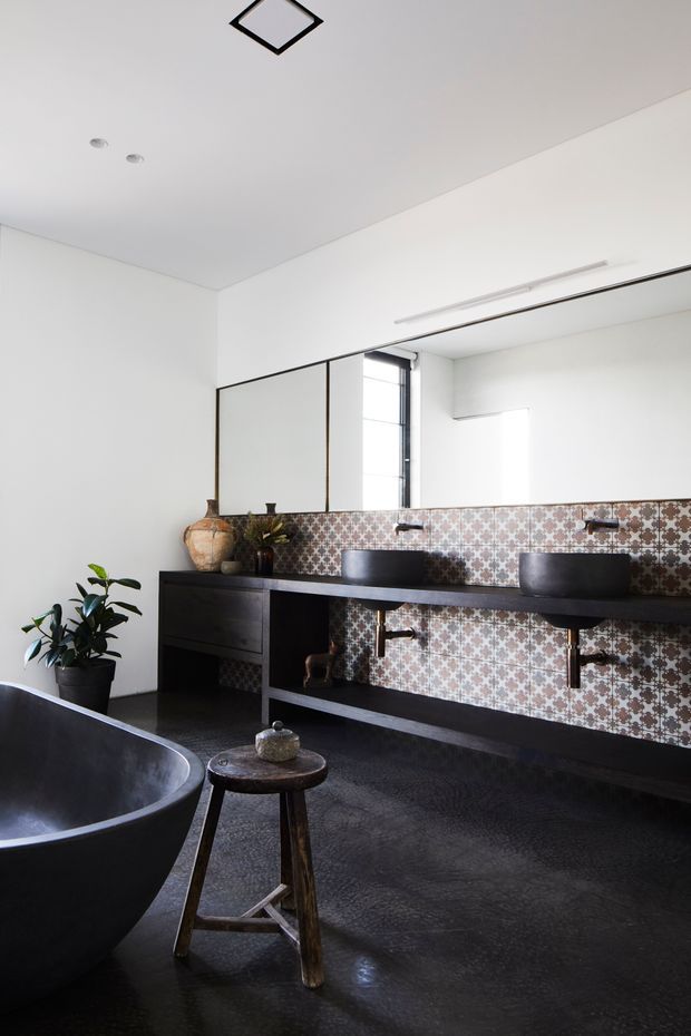 A contemporary bathroom featuring a large freestanding dark bathtub, double vessel basins on a dark built-in vanity, a full-width patterned geometric tile splashback, and a large wall mirror. The room has white walls and ceiling, dark flooring and joinery, black fixtures, recessed lighting, and a potted plant. Natural light enters through a narrow vertical window.