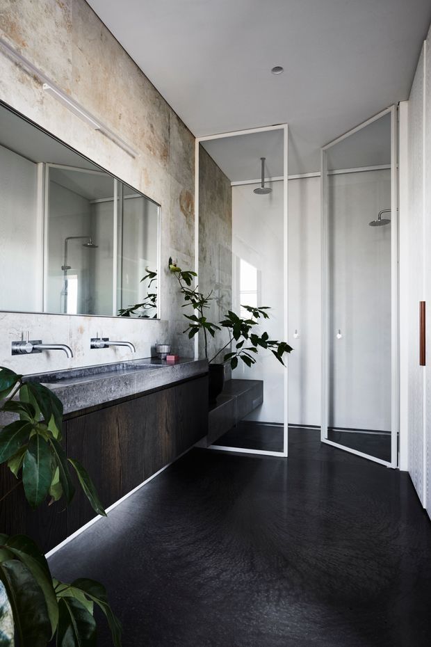 Contemporary bathroom with a long floating dark timber vanity, twin wall-mounted chrome taps and a grey stone benchtop beneath a large mirror. The space includes two glass-enclosed showers with rain showerheads, pale textured wall surfaces, dark flooring, and indoor greenery. Minimal white-framed glazing and clean-lined detailing create a refined, modern atmosphere.