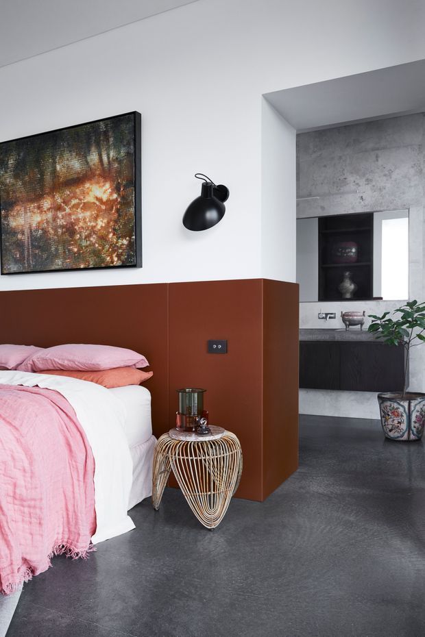 A contemporary bedroom with a partially visible bed dressed in soft pink and white linens, separated from an adjoining bathroom or vanity area by a rust-brown partition wall. The room features dark polished concrete flooring, white walls, a black wall sconce, abstract artwork, and a sculptural wire side table. Through the opening, dark cabinetry, a grey concrete-look wall finish, shelving, and a potted plant are visible.