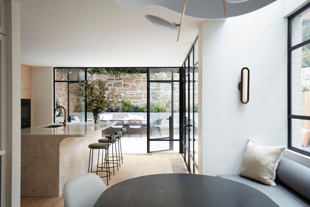 A bright, open-plan contemporary interior combining a kitchen, dining area and living space. A curved stone kitchen island with integrated sink and a row of slim bar stools faces black-framed glazed doors opening to a planted courtyard and exposed stone wall. The foreground includes a dark timber dining table, upholstered seating and a grey sofa beside large black-framed windows. Sculptural ceiling lighting and a slim wall light add refined details.