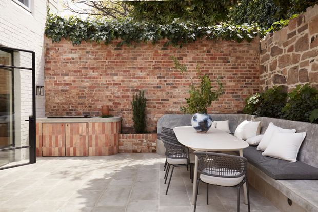 A refined enclosed courtyard dining area with a built-in outdoor kitchen or serving cabinet, pale stone paving, exposed red brick walls, and a built-in upholstered bench. A round dining table with woven chairs sits beneath mature planting and ivy, creating a secluded outdoor entertaining space.