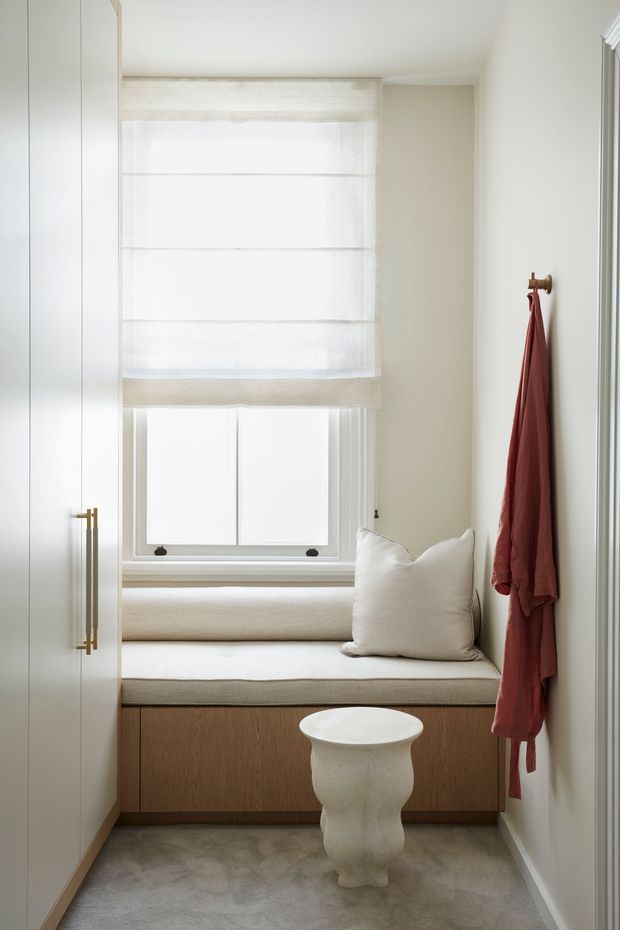 Bright, compact window-seat nook with a built-in upholstered bench, white cushions, pale walls, and full-height white cabinetry with brass handles. A sculptural white side table sits on a soft grey carpet, while a muted terracotta robe hangs from a wall hook. The space is softly lit and refined, with layered Roman blinds over the window.
