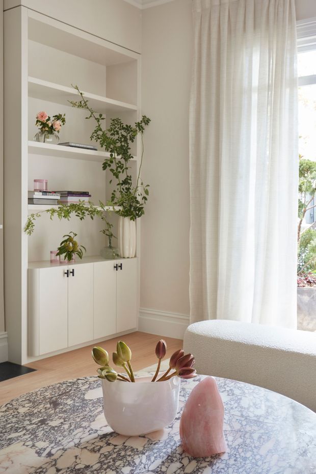 Bright, refined living room corner featuring a built-in white shelving and cabinetry unit, styled with books and trailing plants. Sheer floor-to-ceiling curtains filter daylight beside a window, while a rounded white bouclé chair and a circular marble-patterned coffee table with floral décor complete the scene.