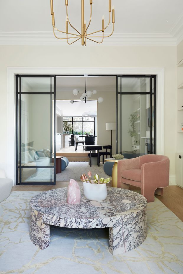 Refined contemporary living room with a sculptural veined stone coffee table, blush upholstered armchairs, pale patterned rug, and a brass linear chandelier. Black-framed glazed steel-look doors open to a connected dining and kitchen area, with layered lighting, decorative joinery, and elegant moulded ceiling details.