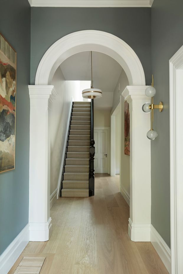A refined residential hallway framed by a large white decorative arch with classical mouldings and columns. The corridor has pale timber flooring, muted blue-grey walls, white trim and skirting, and a carpeted staircase with a dark timber handrail and balusters leading upward. A sculptural pendant light and brass wall sconce add decorative detail, while artwork is visible along the walls.