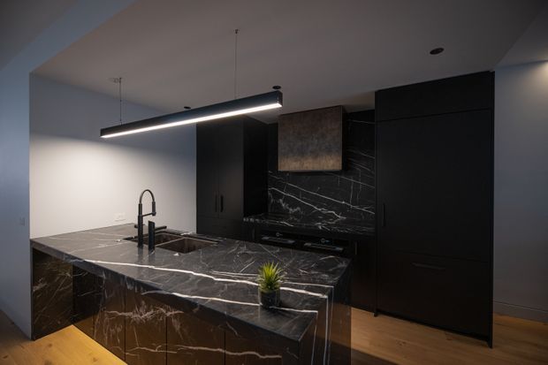 A sophisticated contemporary kitchen with a large dark marble island and matching splashback, black built-in cabinetry, integrated appliances, a black undermount sink and tap, timber flooring, and a linear suspended pendant light. The space has a moody, refined appearance with strong veining in the stone surfaces.