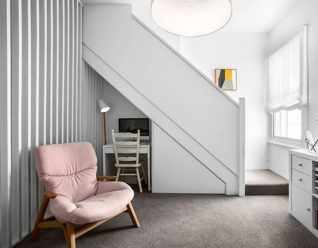 Bright, compact contemporary interior beneath a staircase, featuring a grey carpeted floor, white walls, vertical grey wall panelling, a small desk and chair tucked under the stairs, a pink upholstered armchair, and a white chest of drawers beside a window. The stair enclosure and built-in under-stair workspace dominate the room.