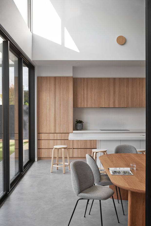 A bright, minimalist open-plan kitchen and dining area with a double-height white ceiling and strong geometric sunlight. Full-height black-framed glazing lines the left side, while warm timber cabinetry spans the rear wall with an integrated white island or breakfast counter. A round timber dining table with upholstered grey chairs and simple timber stools occupies the foreground. The space has a refined, contemporary feel with a polished concrete floor and restrained natural materials.